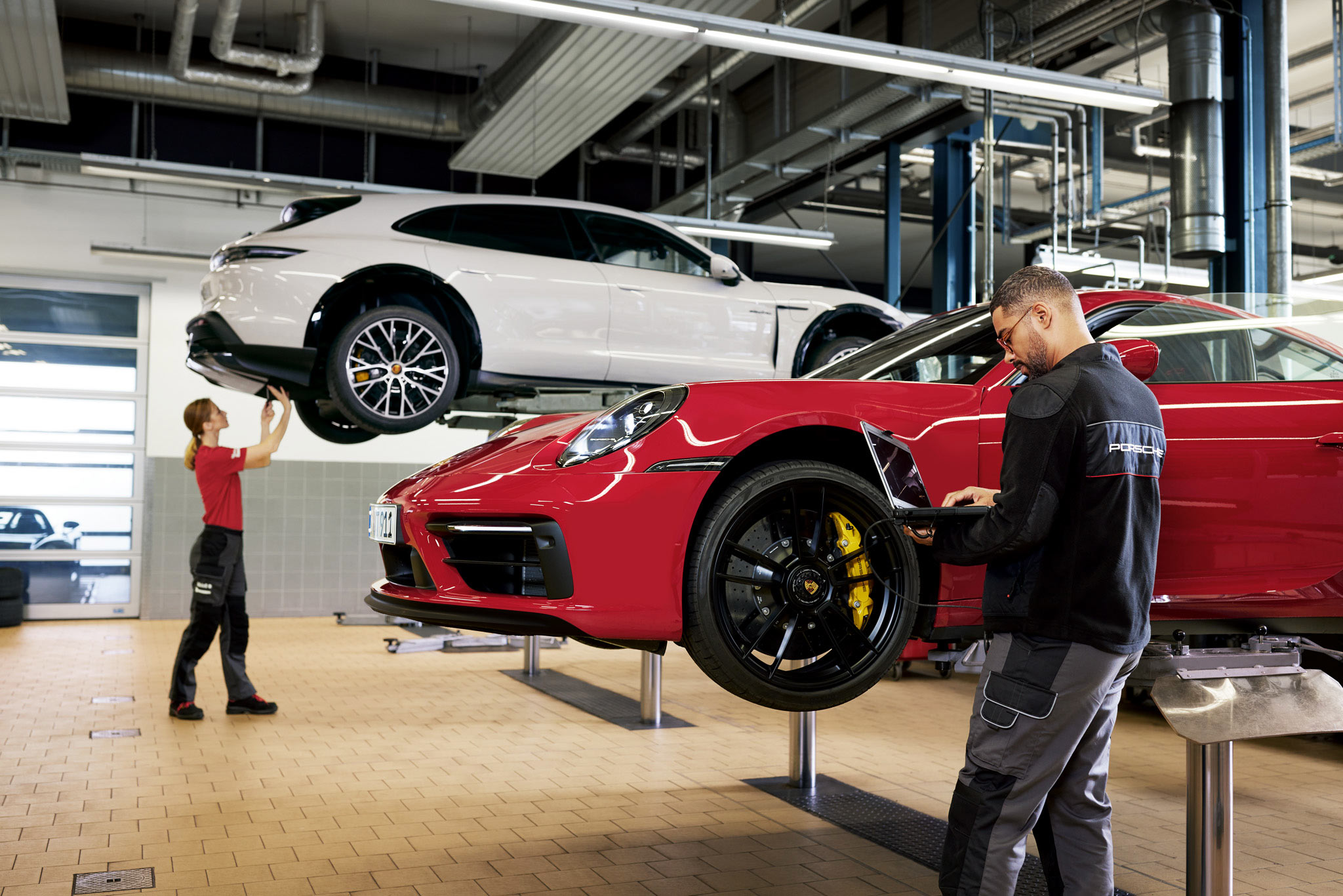 Porsche Service Advisors performing Porsche vehicle maintenance on a white Porsche and a red Porsche in a Porsche Service Center garage