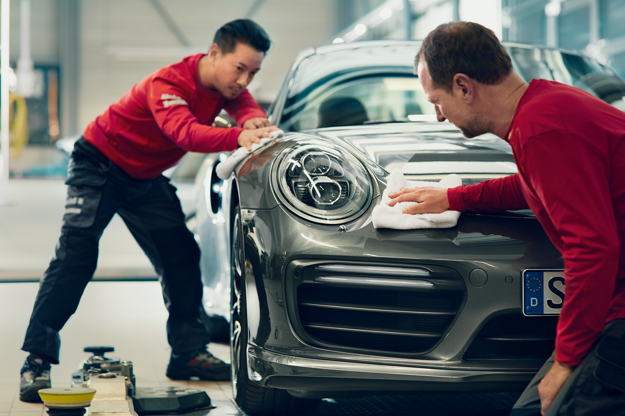 Two technicians meticulously cleaning a gray Porsche in a service center.